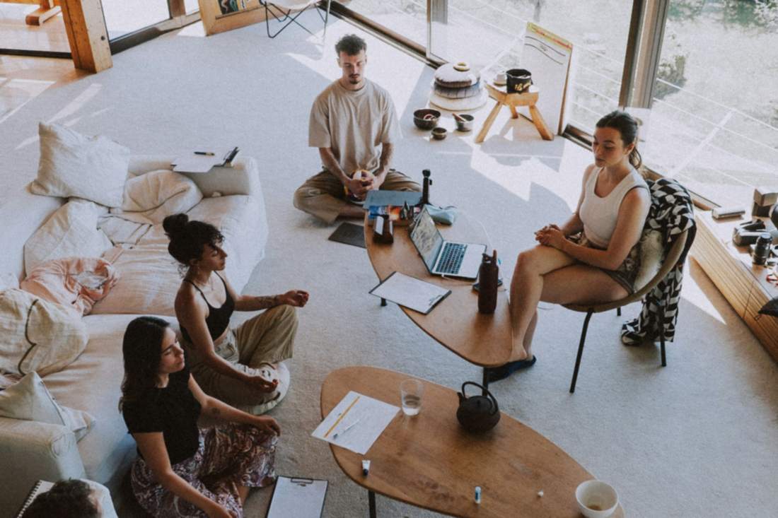 A group sitting together in a bright room, some in meditation, some taking notes.