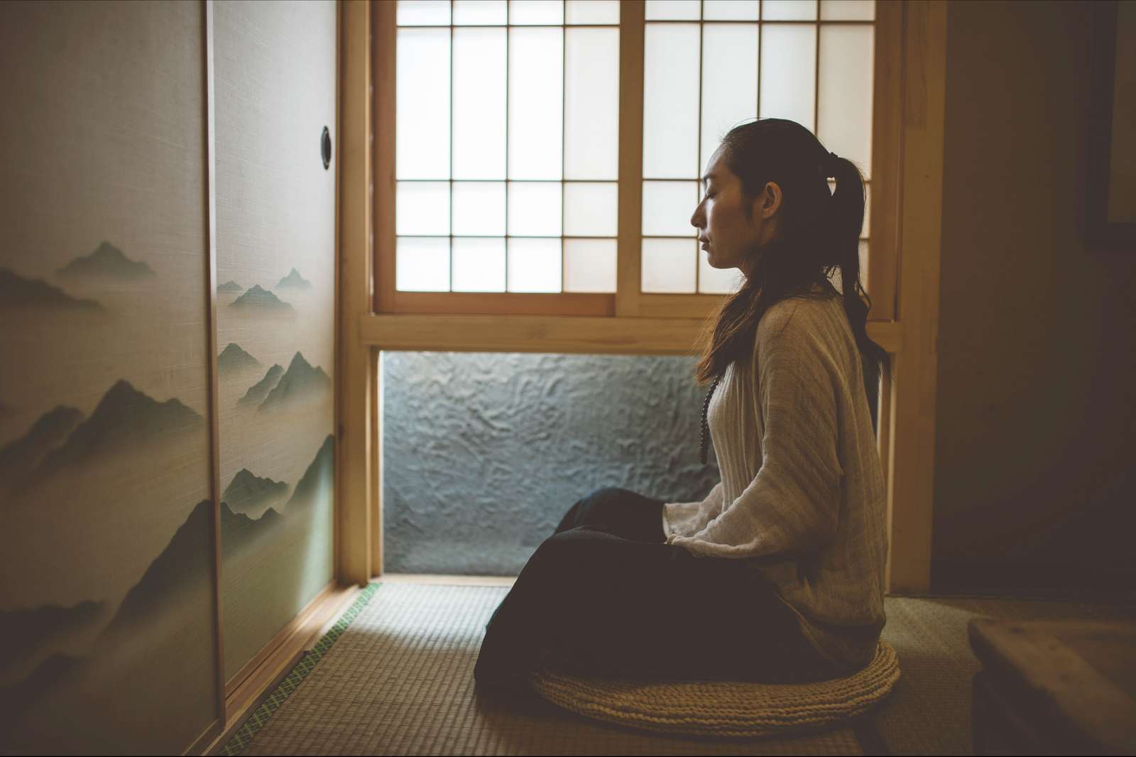 A practitioner sitting on a round cushion on tatami, beside a paper screen and a painted mountain screen.